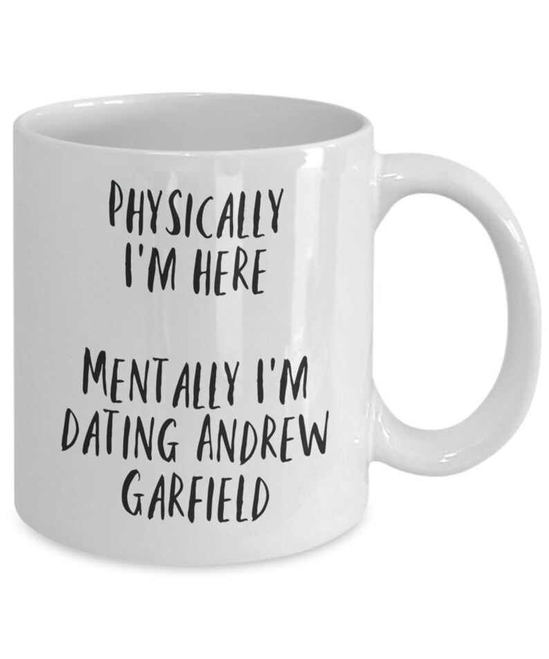 May include: White ceramic mug with the text "PHYSICALLY I'M HERE" above "MENTALLY I'M DATING ANDREW GARFIELD" in black, bold font. The mug has a curved handle and a smooth, glossy finish.