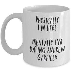 May include: White ceramic mug with a curved handle. The mug features black text that reads "PHYSICALLY I'M HERE" and "MENTALLY I'M DATING ANDREW GARFIELD". The mug is a humorous gift idea.