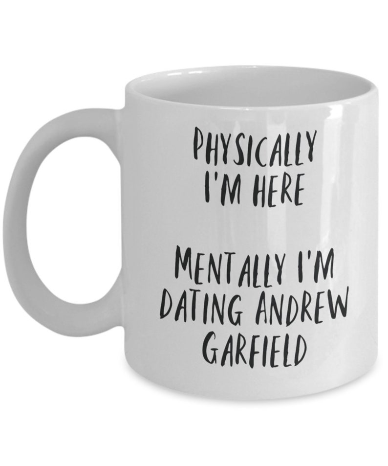 May include: White ceramic mug with a curved handle. The mug features black text that reads "PHYSICALLY I'M HERE" and "MENTALLY I'M DATING ANDREW GARFIELD". The mug is a humorous gift idea.