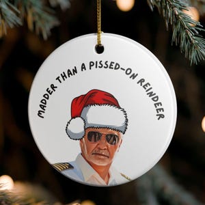 May include: A white, round Christmas ornament with the text "Madder Than A Pissed-On Reindeer." The ornament features a cartoon image of a man wearing a Santa hat and sunglasses.