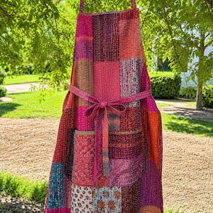 May include: A patchwork apron featuring a vibrant mix of red, pink, orange, and blue fabric squares with intricate kantha-style stitching throughout. The apron includes a front pocket, a waist tie, and a neck strap, all constructed from the same multicolored, patterned textile pieces.