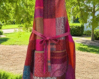 Pode incluir: Um avental de patchwork com uma mistura vibrante de quadrados de tecido vermelhos, rosas, laranjas e azuis com costuras de estilo kantha. O avental inclui um bolso frontal, uma fita para a cintura e uma alça para o pescoço, todos feitos das mesmas peças de tecido multicoloridas e estampadas.