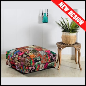 May include: A square patchwork pouf with a vibrant mix of colors and patterns. The pouf is placed on a light wood floor. A small, ornate side table with a plant in a woven basket sits nearby. A turquoise vase hangs on the wall.