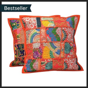 Puede incluir: Dos cojines cuadrados decorativos con un diseño de patchwork. Los cojines presentan una vibrante mezcla de colores, incluyendo rojo, naranja, azul y dorado. Los bordes están ribeteados con una tela naranja lisa. La palabra "Bestseller" está en la esquina superior izquierda.