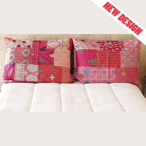 May include: Two rectangular patchwork pillow covers in shades of pink and red, with a variety of patterns and textures. The pillows are placed on a white bed. The words "NEW DESIGN" are in red text in the upper right corner.