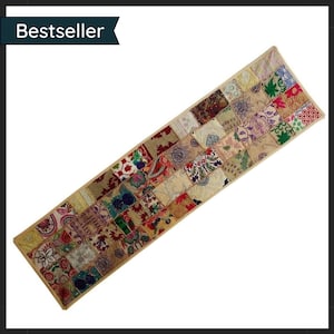 May include: A patchwork table runner with a beige border. The runner is made up of many fabric squares in different colours and patterns, including floral and paisley designs. The text "Bestseller" is in the upper left corner.