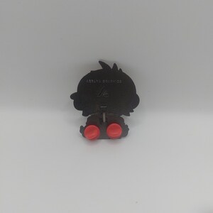 May include: Black metal pin with a cartoon character design. The pin has two red rubber stoppers on the back and the text "AERYA GRAPHICS" is printed on the back.