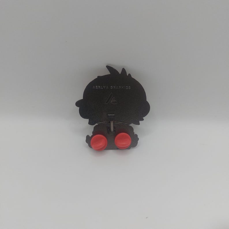 May include: Black metal pin with a cartoon character design. The pin has two red rubber stoppers on the back and the text "AERYA GRAPHICS" is printed on the back.