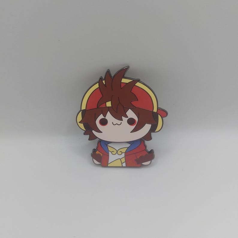 May include: A cartoon character pin with brown hair, a red hat, and a yellow jacket. The character has a cute expression with big eyes and a small mouth.
