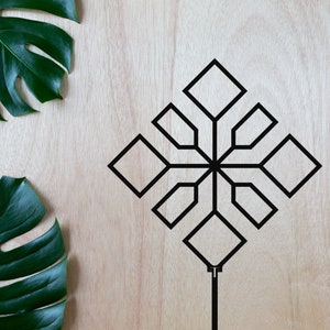 May include: A black geometric design with a stem on a light wood background. Two green monstera leaves are on the left side of the image.