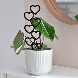 Puede incluir: Una maceta de cerámica blanca con textura acanalada contiene una planta verde con hojas grandes. Una estaca de planta de metal negro en forma de corazón está unida a la planta, con varios corazones apilados uno encima del otro.