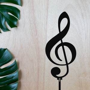 Puede incluir: Un recorte de metal negro de la clave de sol musical sobre un fondo de madera con dos hojas verdes de monstera a cada lado.