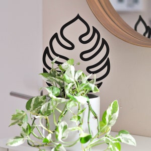 Pode incluir: Um vaso de cerâmica branco com textura canelada contém uma planta em vaso com folhas verdes e brancas variegadas. Atrás da planta, há um recorte de metal preto em forma de folha de monstera.