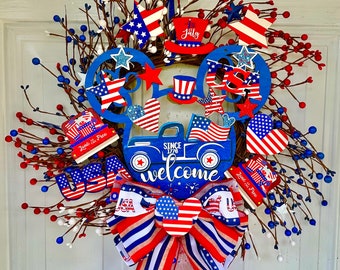 Peut inclure: Une couronne patriotique rouge, blanche et bleue avec une silhouette de Mickey Mouse avec un camion, des étoiles et des cœurs. La couronne est décorée d'un nœud rouge, blanc et bleu et du texte "Bienvenue" et "Depuis 1776".