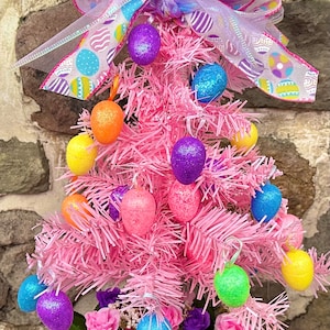 May include: A pink artificial Christmas tree decorated with colorful Easter eggs and a pink and purple bow. The tree is in a white picket fence planter with green and purple flowers.