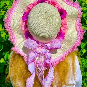 May include: A large, white straw sun hat with a pink and white lace trim. The hat is decorated with pink and purple flowers and a pink ribbon with a floral pattern.