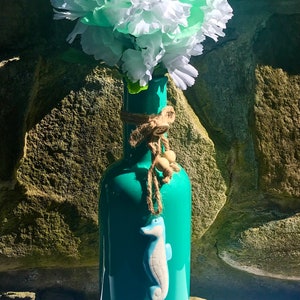May include: A turquoise glass bottle vase with a white seahorse design, decorated with twine and wooden beads. The vase is filled with a bouquet of white and light green carnations.