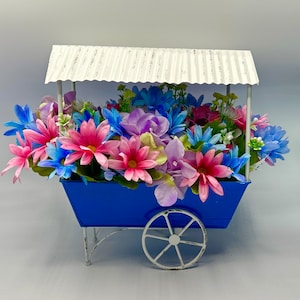 May include: A blue metal wagon with a white roof holds a colorful arrangement of artificial flowers. The flowers are pink, blue, purple, and white.