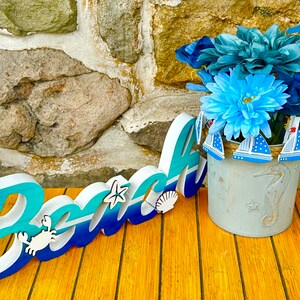 May include: A wooden sign that says "Beach" in blue and white letters with a blue and white seashell, a starfish, and a crab. The sign is sitting on a wooden table next to a metal planter with blue flowers and a seahorse design.
