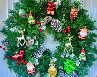 Puede incluir: Una corona de Navidad verde decorada con adornos rojos y dorados, incluyendo un muñeco de nieve, un reno, un pájaro rojo, un oso de peluche, un Papá Noel, un búho y piñas.