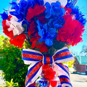 May include: A glass vase filled with red, white, and blue artificial flowers, accented with thin white decorative sticks. A large, multi-layered bow featuring red, white, and blue glittered stripes is tied around the neck of the clear glass vase.