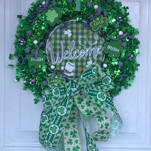 May include: A green and white St. Patrick's Day wreath with a welcome sign, shamrock accents, and a green and white bow. The wreath is decorated with green and white checkered ribbon, green shamrocks, and white beads.