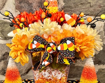May include: A Halloween centerpiece with orange, yellow, and white flowers, a candy corn bow, and a clear vase filled with candy corn. The centerpiece is surrounded by small orange and yellow trees.