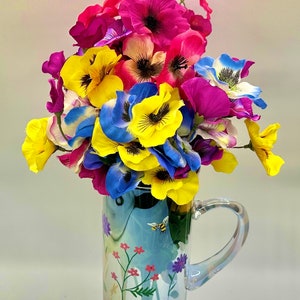 May include: A colorful bouquet of pink, yellow, and blue flowers in a clear glass pitcher with a floral design. The pitcher has a handle and a bee design.