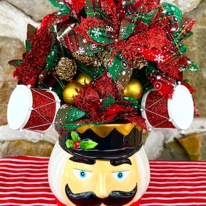 May include: A festive Christmas decoration featuring a ceramic nutcracker head with a black hat and a large mustache. The head is filled with red and green ribbons, gold ornaments, and miniature red drum decorations. The decoration sits on a red and white striped surface.