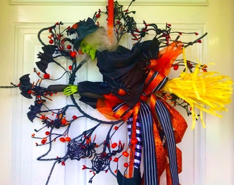 May include: A witch with green skin and a black hat flies on a broomstick with a yellow and orange bow. The witch is surrounded by black bats and spiders on a black wire wreath.