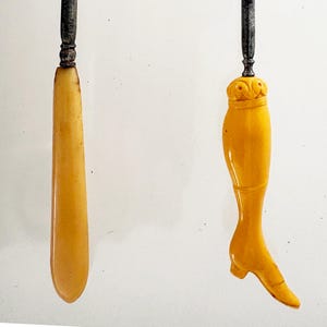 May include: Two antique ivory-colored handles with ornate silver-toned tops. One handle is a long, slender shape. The other is a carved leg with a heeled shoe. The items are likely vintage or antique collectibles.