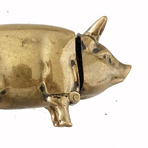 May include: A brass pig-shaped container with a hinged opening. The container is a golden color and has a loop on the back for attaching a chain. The pig has detailed features, including legs, ears, and a snout.