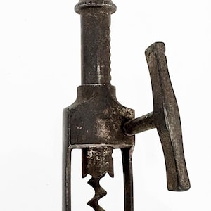 Pode incluir: Um saca-rolhas de metal vintage com um acabamento escuro e desgastado. A ferramenta inclui um cabo em forma de T no topo, um eixo central vertical com mecanismo de parafuso e uma alavanca lateral para operar a espiral de metal.