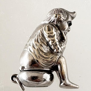 May include: A silver figurine depicting a seated figure, possibly a cherub, in a thoughtful pose. The figure is seated on a small, rounded base with a handle. The details of the clothing and facial features are finely crafted in the silver metal.