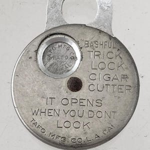 May include: A silver metal cigar cutter with a ring on top. The cutter has the text "Bashful Trick Lock Cigar Cutter" and "It opens when you don't look" on it. The text "TAFO MFG. CO. L.A. CAL" is also on the cutter.
