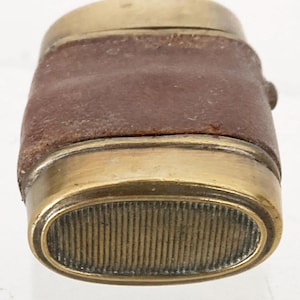 Op de afbeelding: Een vintage aansteker met een bruine leren wikkel en messing accenten. De aansteker heeft een ovale, getextureerde basis en een messing bovenkant. Het leer vertoont tekenen van slijtage, wat duidt op leeftijd en gebruik. Het ontwerp is compact.