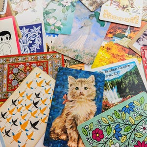 May include: A collection of vintage postcards and playing cards featuring diverse illustrations including a fluffy kitten, a cat, floral patterns, a landscape of The Butchart Gardens, a cartoon couple with the text Love is, and a girl holding cards.