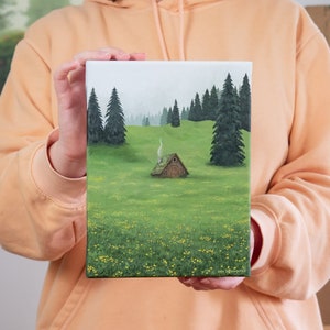 Può includere: Un piccolo cottage marrone con una colonna di fumo che sale dal camino si trova in un campo verde con fiori gialli selvatici. Il cottage è circondato da una foresta di alberi verde scuro.