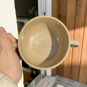 Op de afbeelding: Een beige keramische kom met twee handvatten, van bovenaf gezien. De woorden "Thank You" staan in de kom gegraveerd. De kom wordt in de hand van een persoon gehouden, met een gestreept tapijt op de achtergrond.