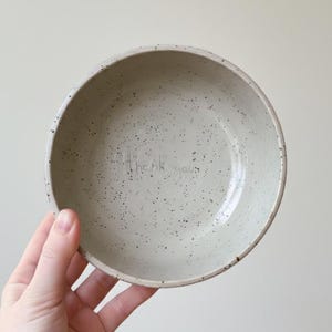 Puede incluir: Un cuenco de cerámica moteada en un color neutro se sostiene en una mano. El cuenco tiene una textura ligeramente rugosa y las palabras "thank you" están escritas en el centro. El cuenco es de color beige claro con pequeñas motas negras.