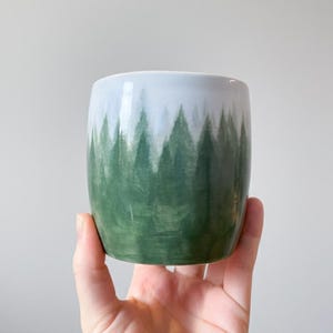May include: A ceramic tumbler with a forest design. The cup is cylindrical with a white top and a green forest painted on the lower half. The forest design features stylized evergreen trees. The cup is held in a hand.