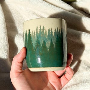 May include: A ceramic tumbler with a forest scene painted around the exterior. The forest is depicted in shades of green, with a cream-colored top. The tumbler is held in a hand, with other ceramic pieces in the background.