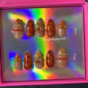 May include: A set of ten artificial nails in a pink box. The nails feature a variety of designs, including a nude base with brown marble effect, gold rings, and gold accents. The text "slayedbykymlee" is visible on the box.