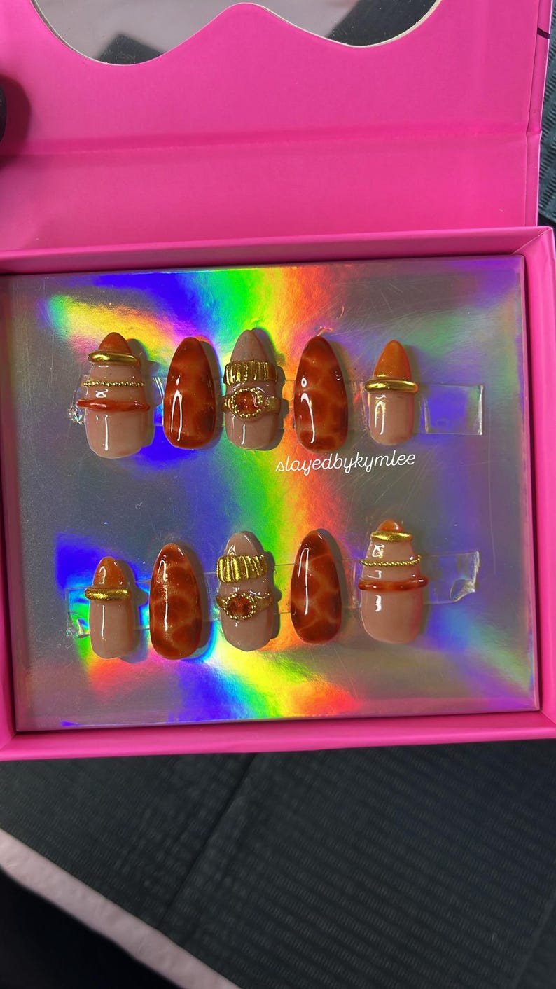 May include: A set of ten artificial nails in a pink box. The nails feature a variety of designs, including a nude base with brown marble effect, gold rings, and gold accents. The text "slayedbykymlee" is visible on the box.