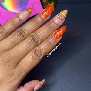 May include: Close-up of a hand with long, almond-shaped nails. The nails are painted in a combination of nude and vibrant orange shades, with gold accents. The text "slayedbykymlee" is visible on the hand.