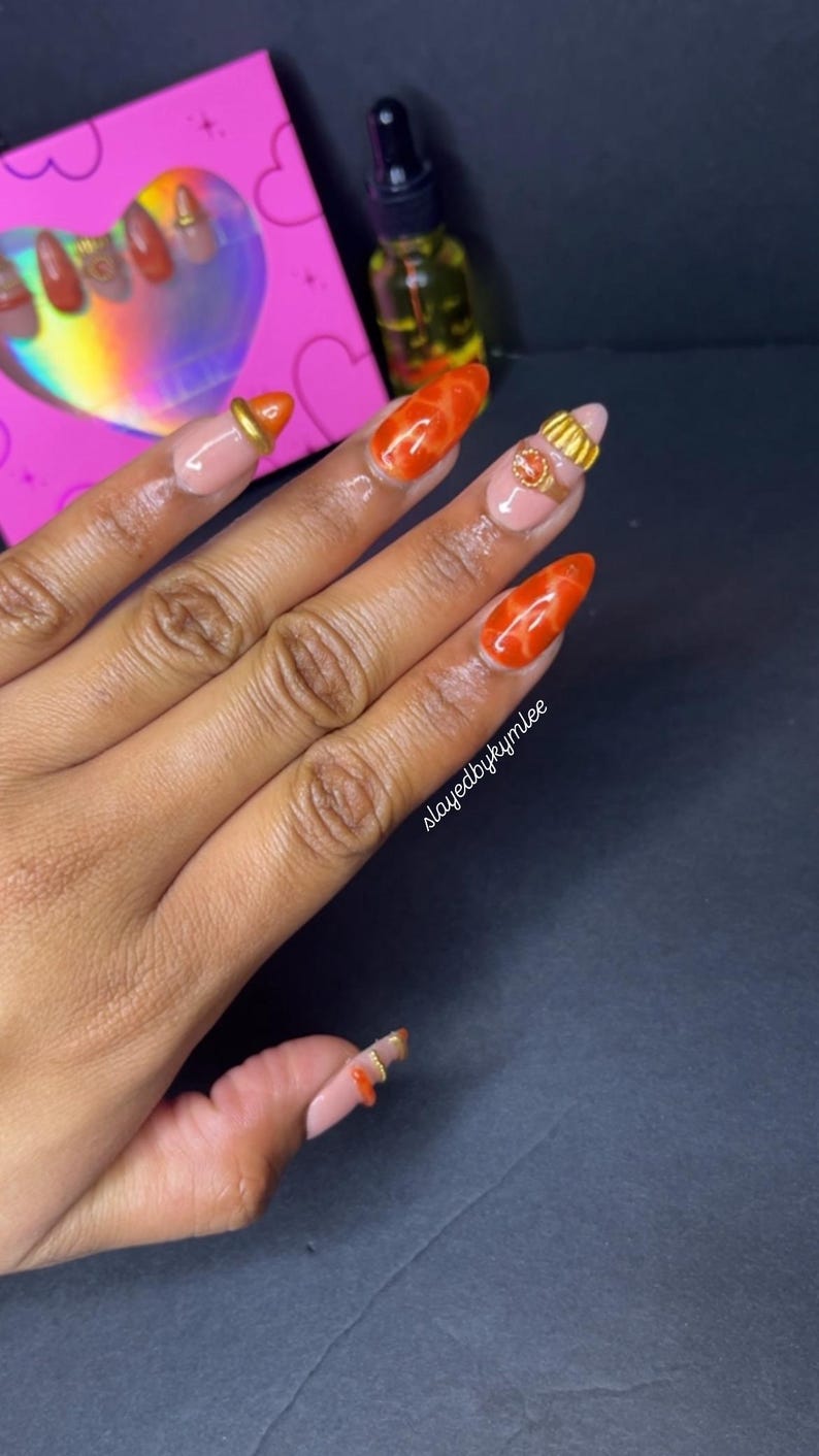 May include: Close-up of a hand with long, almond-shaped nails. The nails are painted in a combination of nude and vibrant orange shades, with gold accents. The text "slayedbykymlee" is visible on the hand.