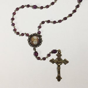 Puede incluir: Un rosario de cuentas de vidrio morado con un crucifijo de latón y un medallón redondo con el retrato de un hombre.