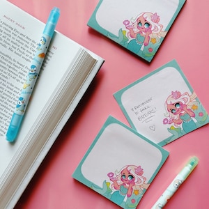 May include: A collection of stationery items on a pink surface. Includes a blue highlighter pen with a cartoon penguin design, a book, and several square sticky notes with mermaid illustrations and the text "Remember to take BREAKS!"