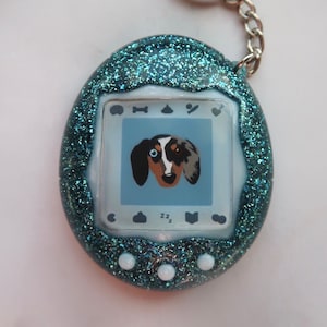May include: A teal blue glitter Tamagotchi with a white screen displaying a cartoon dog. The screen also shows icons for food, poop, a game, and a heart. The Tamagotchi has a silver keychain attached.
