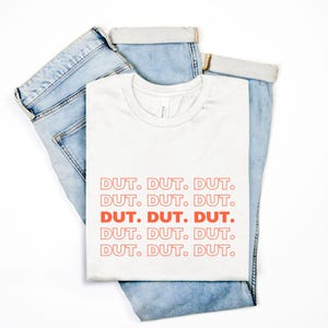 May include: A white t-shirt with the word "DUT" repeated in a stacked pattern in orange. The shirt is laid on top of a pair of light blue jeans.